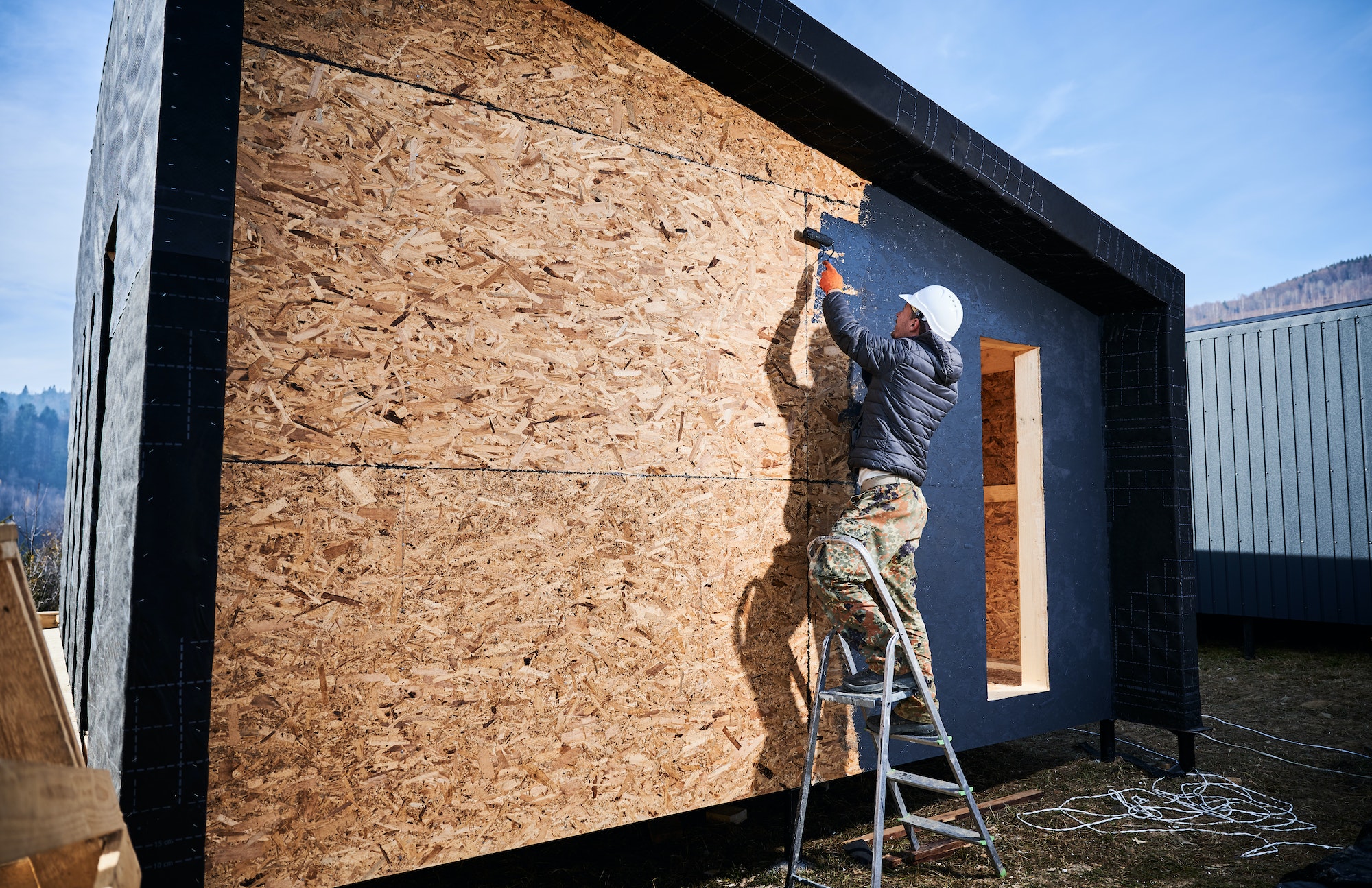 Male painter using paint roller, doing exterior paint work while building wooden frame house.