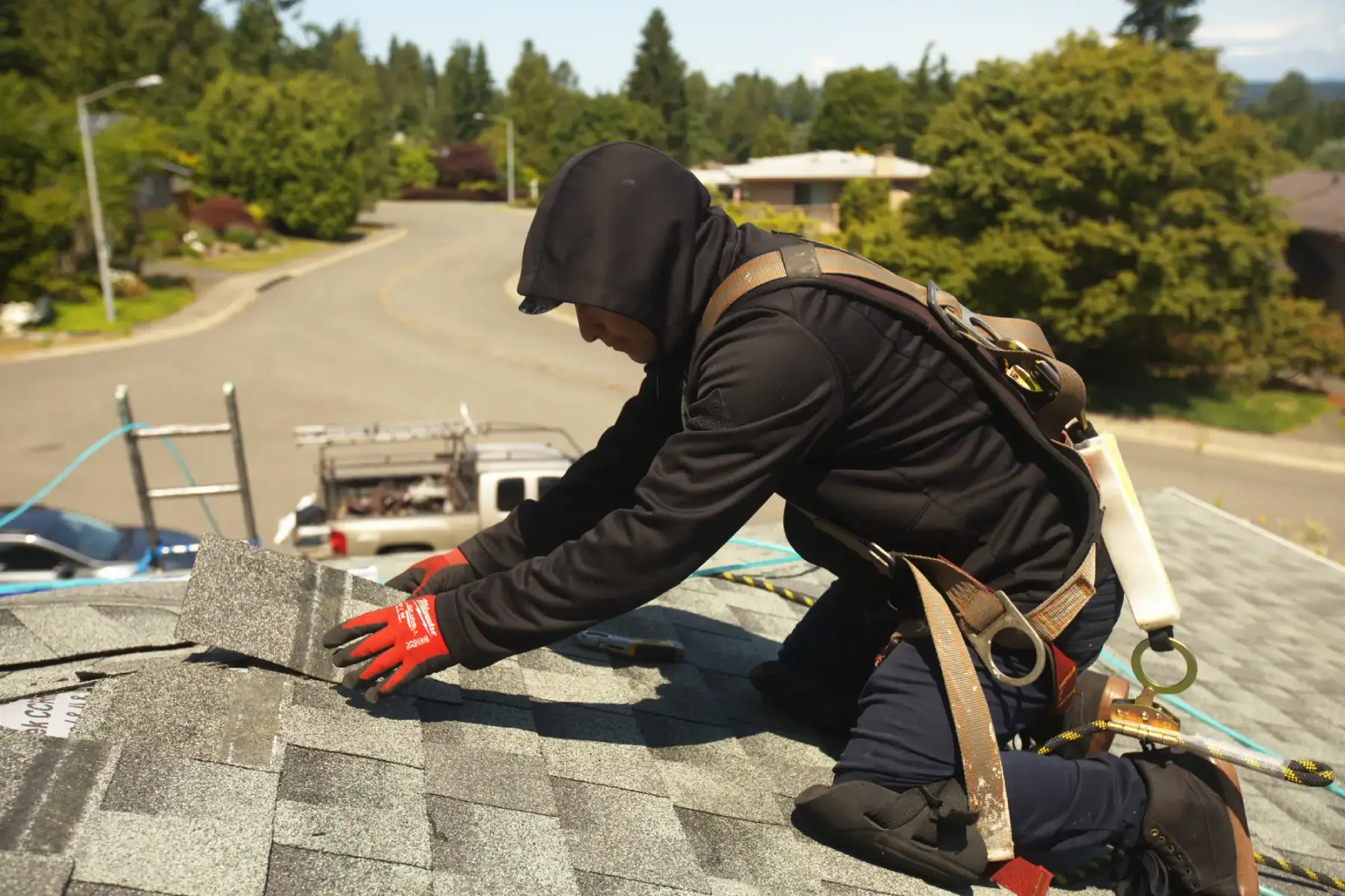 Roofer installing shingles for residential roofing services while wearing safety harness on a sunny day.